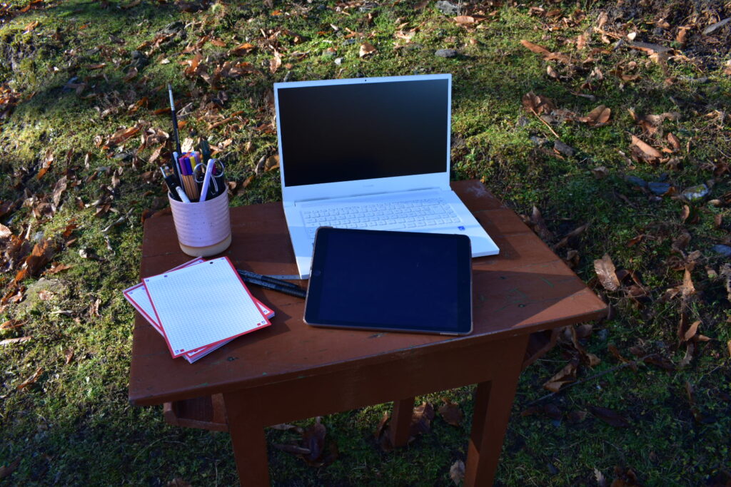 Bureau de travail en extérieur avec ordinateur portable, carnet et outils numériques, posé dans un environnement naturel.